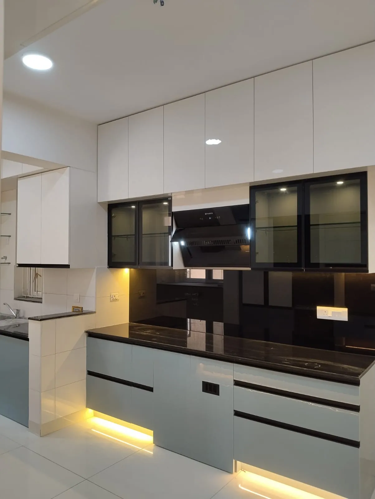 Modular kitchen with white upper units, black-framed glass shutters, a chimney, black glass backsplash and grey base drawers with LED lighting, House Decors, Chennai