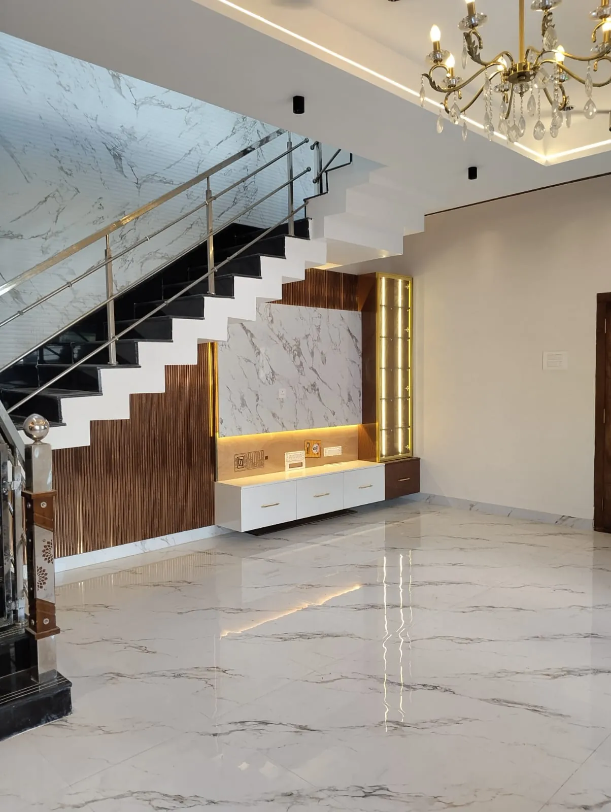 Living area with a staircase, marble-look wall, wooden slat panelling and a wall-hung unit beside a lit glass display column, House Decors, Chennai
