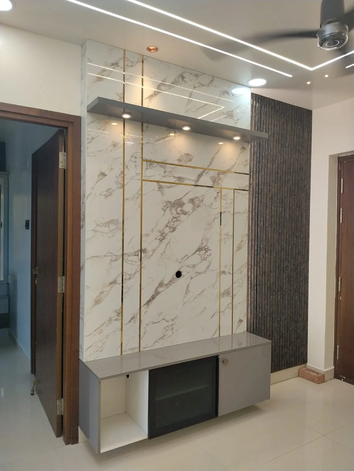 TV unit with a marble-look panel, gold strips, a floating shelf with spot lights and a dark fluted wall panel, House Decors, Chennai