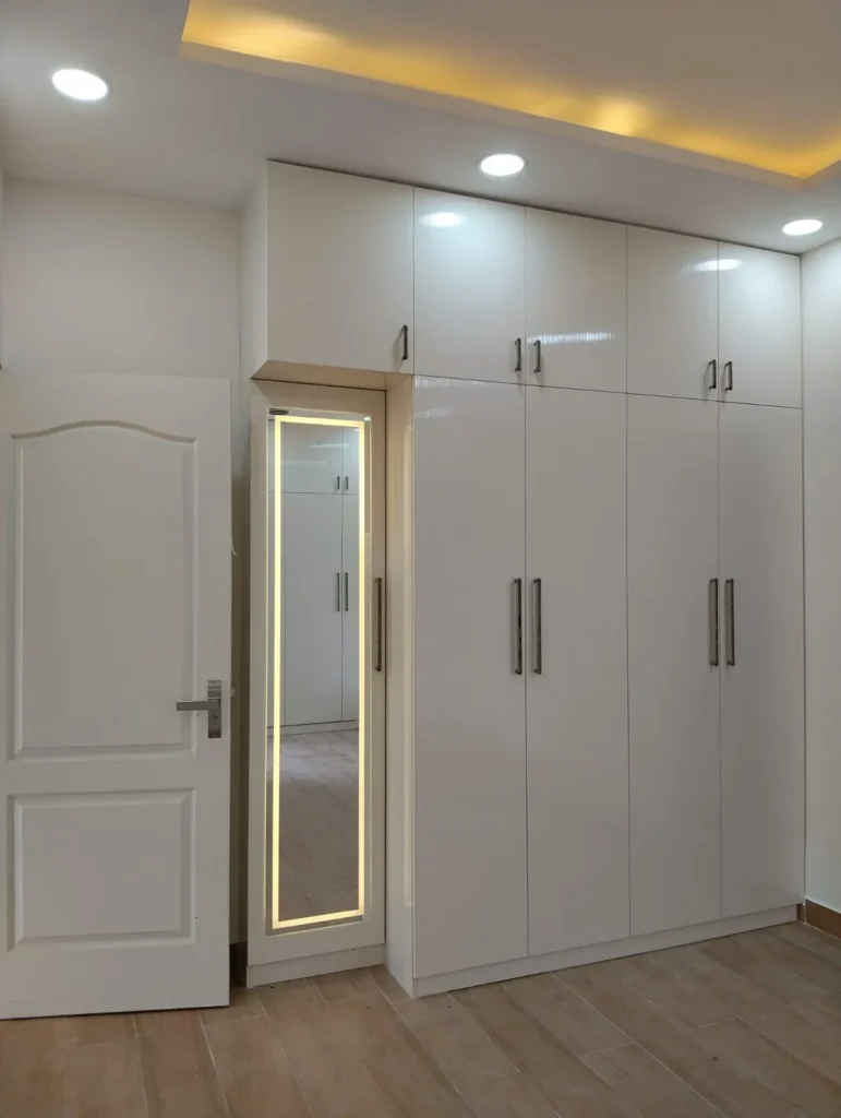 White wardrobe with loft and an LED-framed full-length mirror beside a white door, House Decors, Chennai